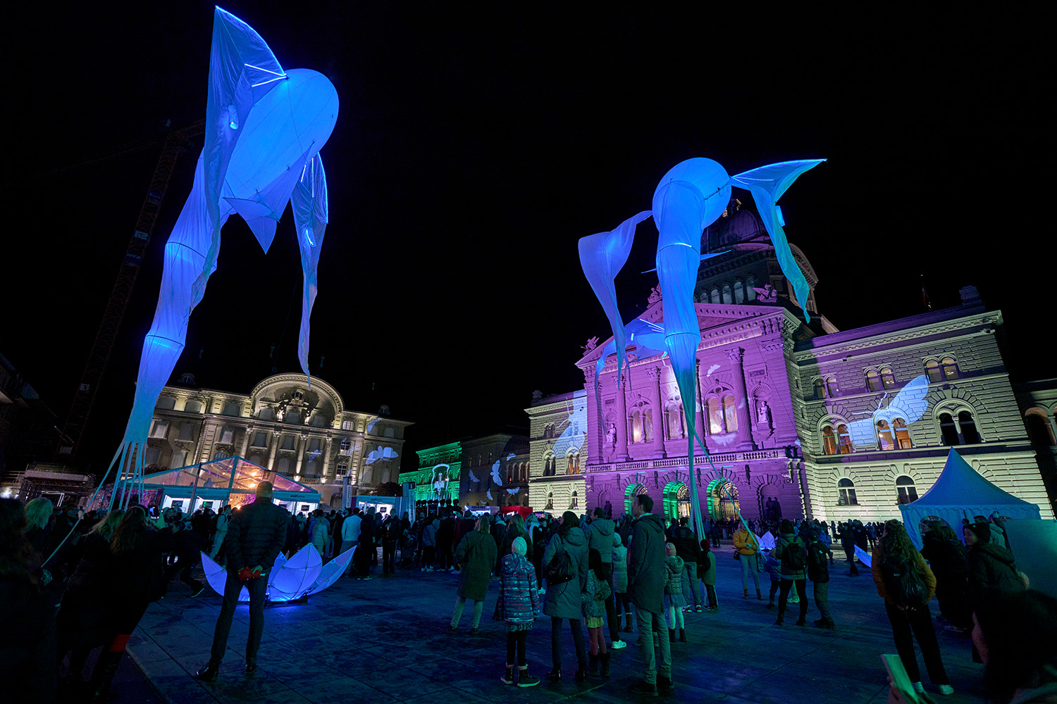 Der Bundesplatz an der Museumsnacht mit dem farbig beleuchteten Bundeshaus und der Nationalbank. Auf dem Platz schauen viele Menschen den zwei «Luminéoles» zu. Diese Fantasiewesen aus Licht, tanzen mit ihren grazilen Flügeln im Wind.